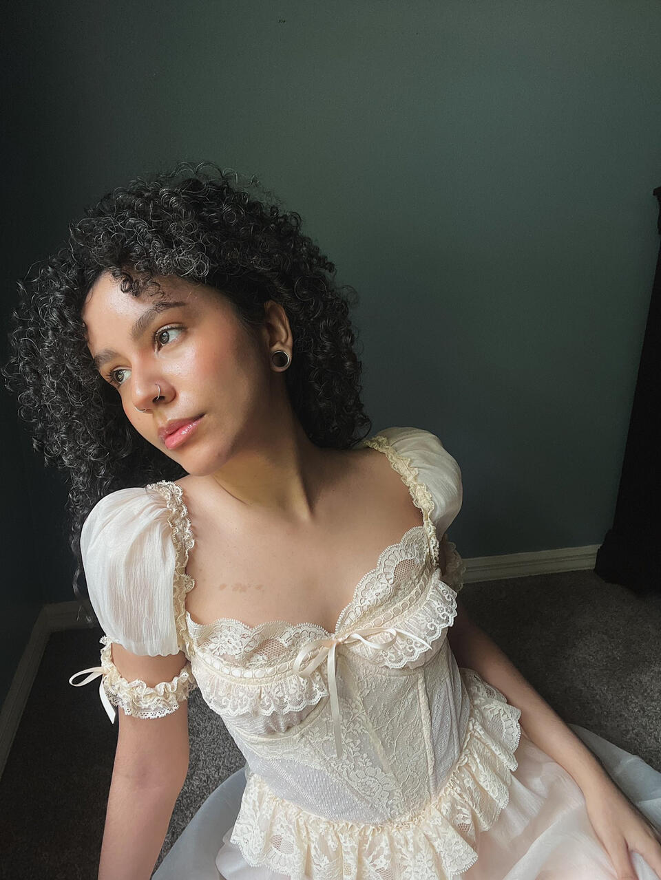 Sopa is wearing a lacey, puff sleeve, corseted dress. She’s staring off in the distance at a 3/4 profile. She has brown eyes, black curly hair, and neutral light olive toned skin.